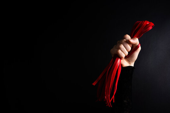 Human hand holding red flogger whip against dark background, studio lighting highlighting leather straps, texture, and grip.