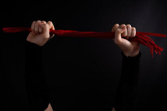 Human hands holding red flogger whip stretched horizontally against dark background, studio lighting highlighting texture and grip.