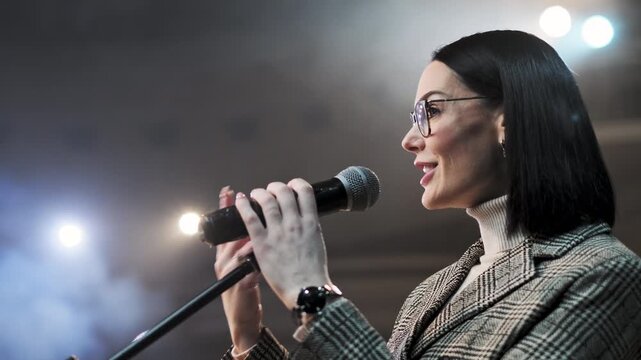 Confident businesswoman in glasses speaking into microphone while addressing audience on stage. Female presenter delivering speech at conference, seminar or corporate event under professional lighting