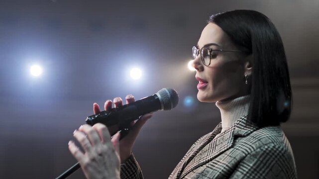 Professional woman in glasses speaking into microphone and gesturing while presenting on stage under bright lights. Female speaker delivering speech, leading seminar or hosting business event. Concept