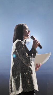 Confident businesswoman in glasses speaking into microphone while addressing audience on stage. Female presenter delivering speech at conference, seminar or corporate event under professional lighting