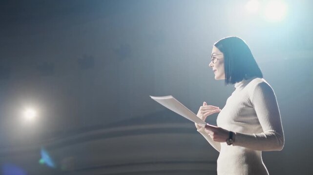 Professional business woman speaking on stage while holding notes under bright spotlight. Female presenter gesturing during speech, delivering presentation or keynote in corporate environment. Concept