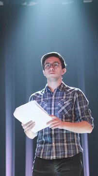 Focused young man holding script and looking up under dramatic spotlight on stage. Actor reading lines, preparing for audition or rehearsing performance in dark theater environment with curtains