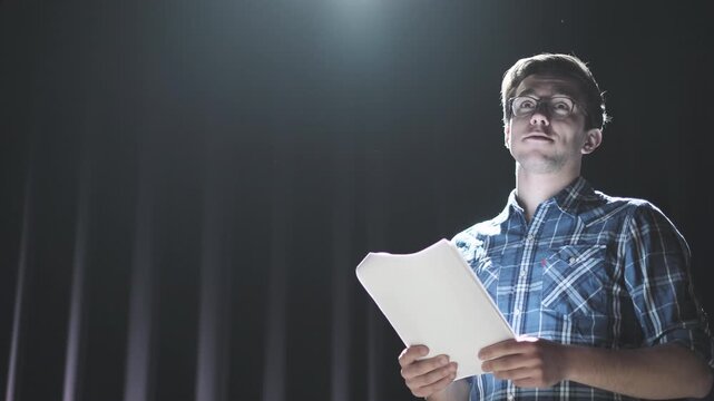 Focused young man standing on stage under bright spotlight holding script and gesturing while speaking. Actor rehearsing lines or practicing performance in theater setting with dramatic lighting and