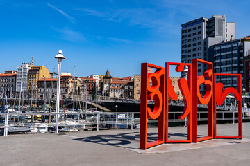 The famous red "Gijón" sculpture, known as Las Letronas, stands at the city's marina with sailboats and historic buildings in the background under a clear sky. © cimata