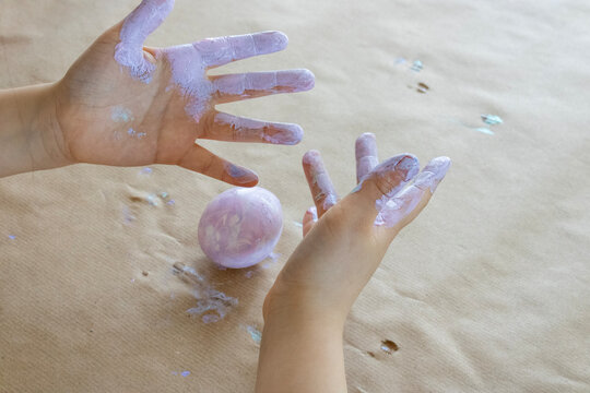 Messy hands of a young child covered in purple pastel paint, holding a decorated Easter egg. Top view of a sensory art play and creative mess on a brown kraft paper background. Concept of childhood jo