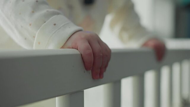 Close-up of infant girl hand resting on crib railing while standing in baby bed. Early childhood development and first support skills at home