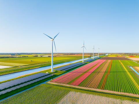 Aerial view of Dutch countryside with wind turbines and solar panels among green fields. Sustainable energy and agriculture in Netherlands under blue sky. Summer sunny day rural landscape from above.