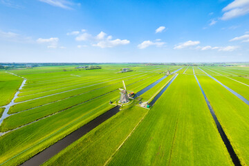 Naklejka premium Aerial view of traditional Dutch windmills by water canals in a green field. Historic heritage site in Netherlands under a blue sky. Rural landscape with classic architecture and agriculture.