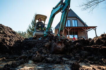 Fototapeta premium A person operates a teal mini excavator, digging wet soil before an A frame cabin with large vertical windows and a timber porch, conifers border the rural site.