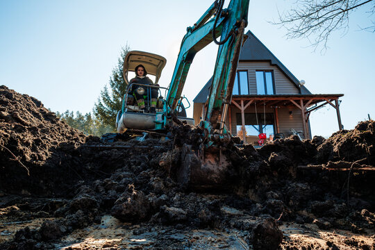 A person operates a teal mini excavator, digging wet soil before an A frame cabin with large vertical windows and a timber porch, conifers border the rural site.