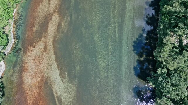 Po?itelj, Bosnia and Herzegovina - 05 17 2025: Top-down drone video of Neretva River, showing sandy riverbed, deep green waters, lush trees along the banks, and abstract water patterns on a sunny day.