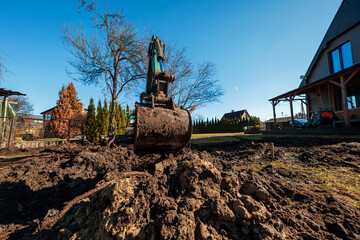A compact excavator with a mud caked bucket digs churned soil in a suburban yard, framed by leafless trees, a conifer hedge, and wooden houses with steep roofs. © True Pixel Art