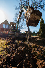 Fototapeta premium A homeowner runs a compact excavator, lifting wet soil beside a wooden gable roof house with large vertical windows, in a yard bordered by conifers on a bright late afternoon.