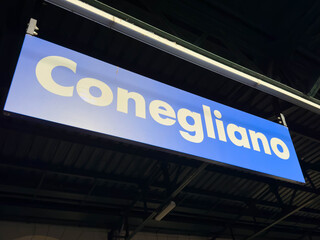 Fototapeta premium Conegliano, Italy - January 27, 2026: Blue sign displaying the name of the train station Conegliano, mounted on a metal structure with overhead lighting visible in the background