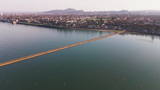 Cinematic aerial flyover along the historic Granton pier towards the Edinburgh skyline and coastal beach, Scotland