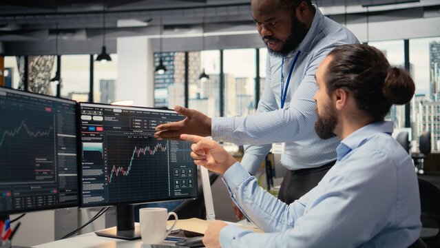 Prop firm staff members communicate at PC desk to ensure smooth trading operations. Team of traders in office doing brainstorming, discussing market trends and portfolio positions