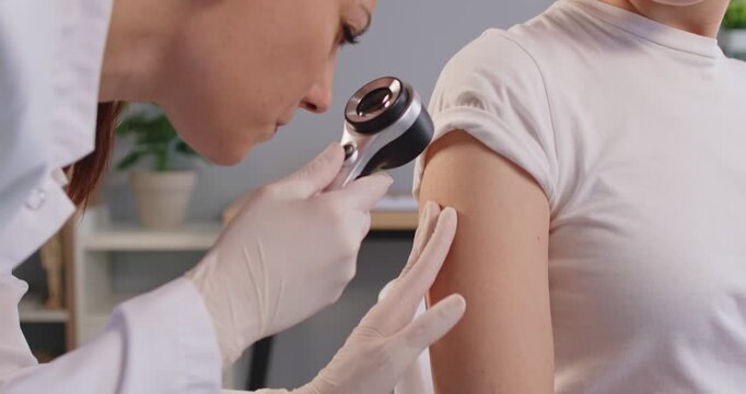 Close up of female dermatologist using dermatoscope and closely examining patient skin on upper arm. Medical professional performing dermatological checkup to assess mole or skin condition in clinic.