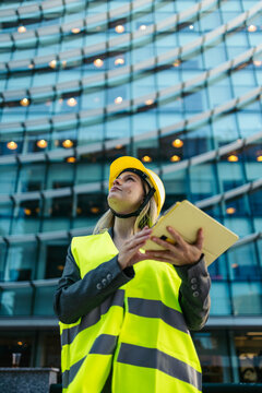 Woman architect wearing hardhat and safety vest holding tablet, looking up at building