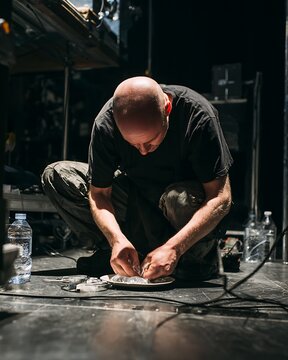 A man crouches attentively over a surface, with bottles and wires scattered around him. He appears to be deeply focused on his task, bathed in dim light