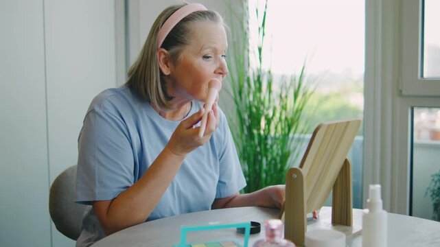 Elderly woman applies makeup in bright room with plants and a vanity mirror during late morning