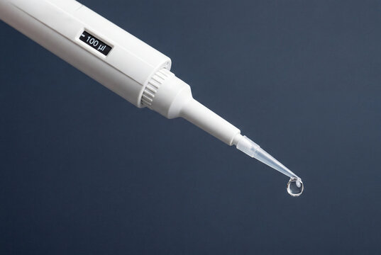 White medical pipette holding a single clear liquid drop at the dispensing tip over a dark surface