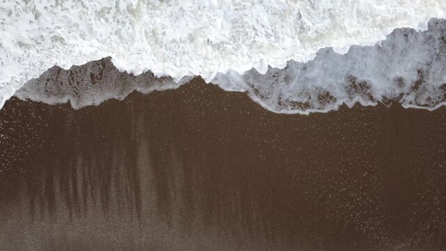 Static aerial top-down view of ocean waves crashing on a sandy beach. Detailed abstract composition suitable for both horizontal and vertical use. Long background footage. 4K