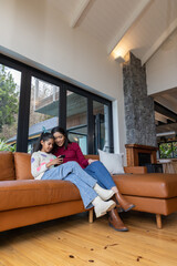 Fototapeta premium Asian mother and child sitting in living room on cognac sofa near glass doors, using smartphone