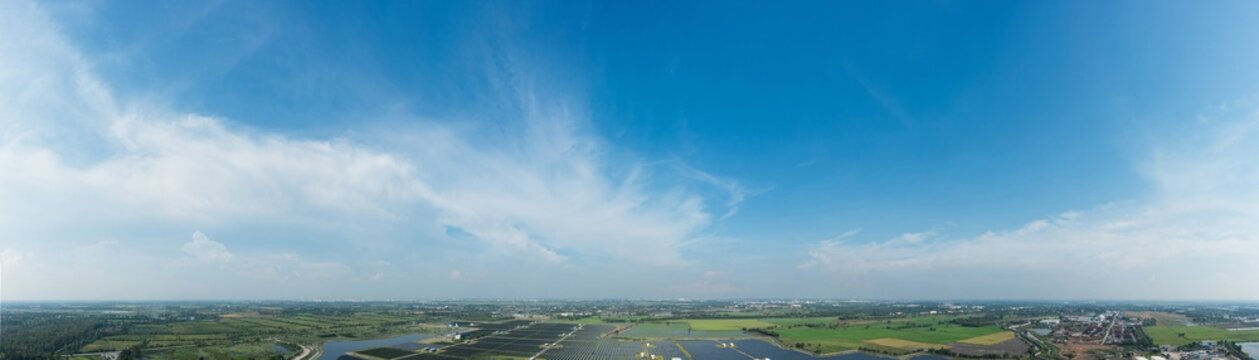 Panoramic wide-angle view blue sky with soft clouds over green rural landscape and distant industrial area, showcasing environmental balance, sustainability and clean energy concept with natural light