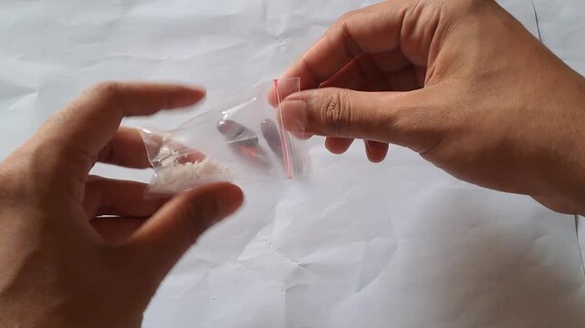 Close-up of Asian male hands demonstrating the dangers of illegal drugs using capsules and a small plastic bag of white powder as visual aids for drug abuse prevention education.