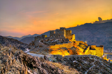 A scenic view of the ancient stone structures of Thee Ain Heritage Village illuminated at sunset against the mountains in Al Bahah, Saudi Arabia.