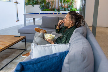 Naklejka premium African American male sitting on gray sectional holding bowl of popcorn in lounge near pool table