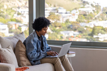 Fototapeta premium African American adult man sitting on beige sofa at home wearing denim jacket, using laptop