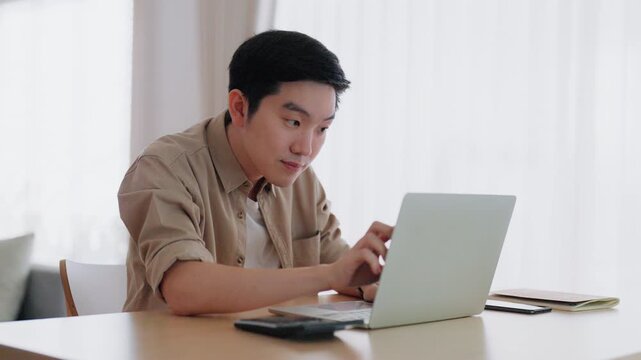 Happy young Asian man working on laptop in living room. Freelance worker male sitting at desk writing notes while watching webinar, studying online