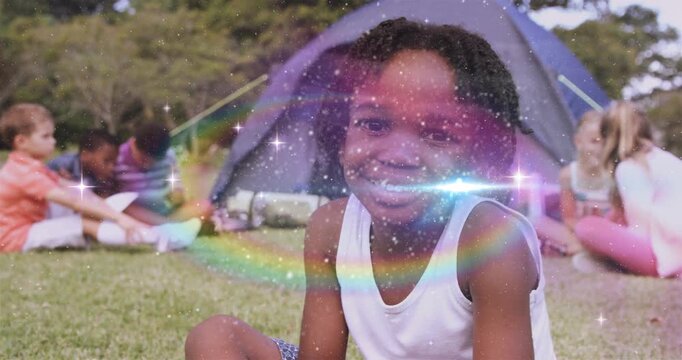 Front girl sitting on grass smiling, sparkles emerging, rainbow sweeping face, enhancing camping