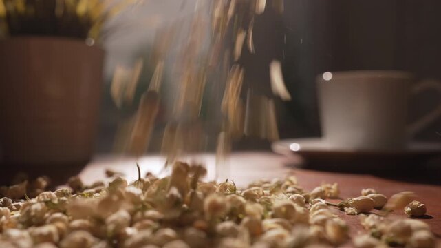 Cinematic shot of dried jasmine buds pouring down continuously onto a prep table in warm natural sunlight, with a delicate tea cup in the background for herbal tea concept.