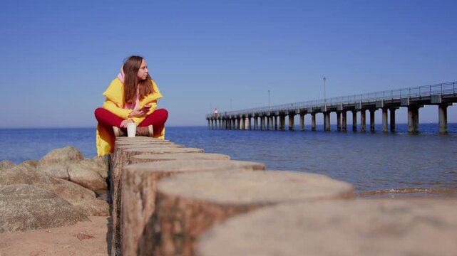 A woman sitting on a wooden breakwater by the sea. A girl in a yellow jacket is resting by the pier, holding a smartphone and a cup of coffee. The theme of recreation, travel, and vacation lifestyle.