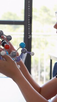 Vertical video: Teen lifting molecule model by window rotating while binary overlay aiding STEM