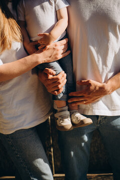 The image shows two adults and a small child. The child is between them, supported by the adults' arms. Only parts of their bodies arms, legs, and clothing are visible, not their faces.

