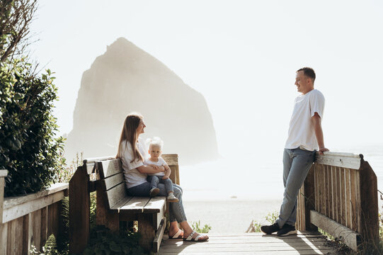 The photograph depicts a family relaxing on a wooden bench by the seashore. The woman sits on a bench, holding a small child, and the man stands opposite, leaning on the railing. They look at each oth