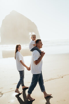 The image shows a family walking along a sandy beach by the sea. The man carries a small child on his shoulders, supporting him with his arms. The child sits quietly and looks forward.