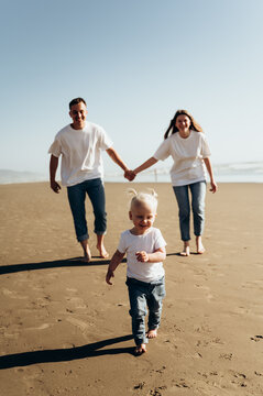 A small child joyfully runs along the beach, leaving barefoot footprints in the sand. His face glows with happiness and curiosity. Behind him, his parents walk hand in hand, smiling and gazing lovingl