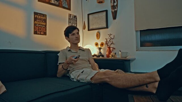 Young man sitting comfortably on the sofa in his living room. He is holding a remote control and changing channels while watching a movie