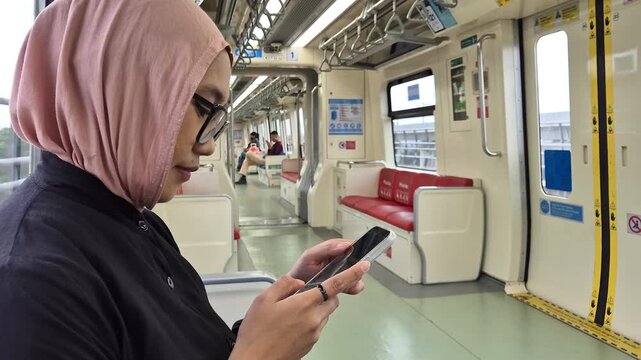 Side view of a young Asian Muslim woman in a pink hijab and glasses using a smartphone while sitting in a modern subway train carriage
