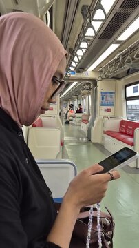 Side view of a young Asian Muslim woman in a pink hijab and glasses using a smartphone while sitting in a modern subway train carriage