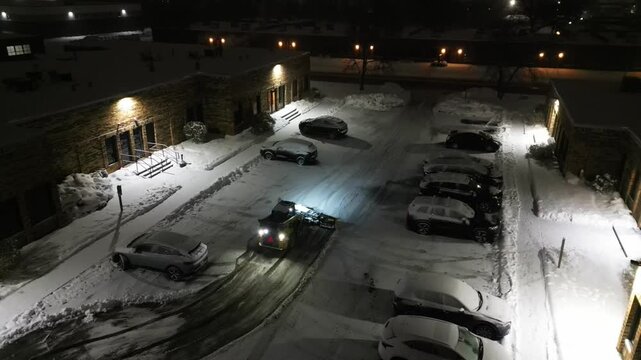 A skid steer loader with a snowplow attachment clears heavy snow from a commercial building's parking lot during a cold winter night.