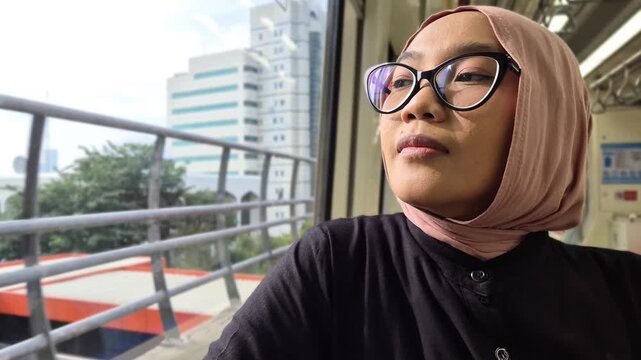 Young Asian Muslim woman wearing a pink hijab and glasses, looking thoughtfully out the window while commuting on a modern city train