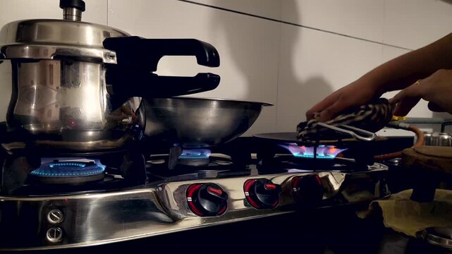 Person cooking on gas stove with blue flame, steel utensils and pressure cooker in home kitchen. Useful for household, food preparation and daily lifestyle content.