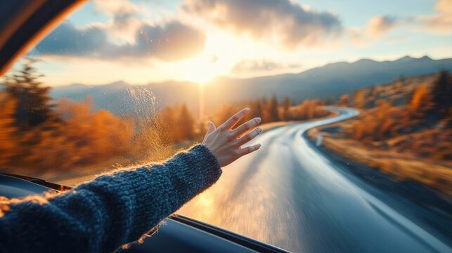 outstretched hand from a car window reaching toward sunset over a winding mountain road lined with autumn trees, motion blur and warm golden-hour freedom