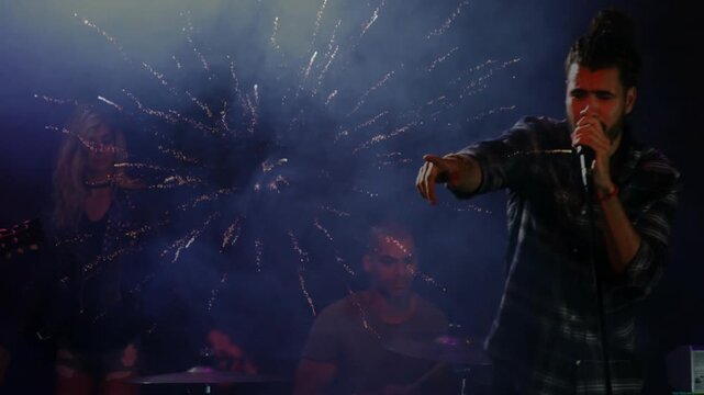 Music singer raising hand after pyro spark burst, moving center, singing into mic, engaging crowd